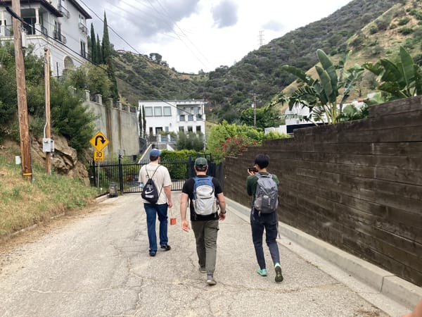 Three people with backpacks walk down a paved trail toward a gate. Behind the gate are a large white mansion and green hills.